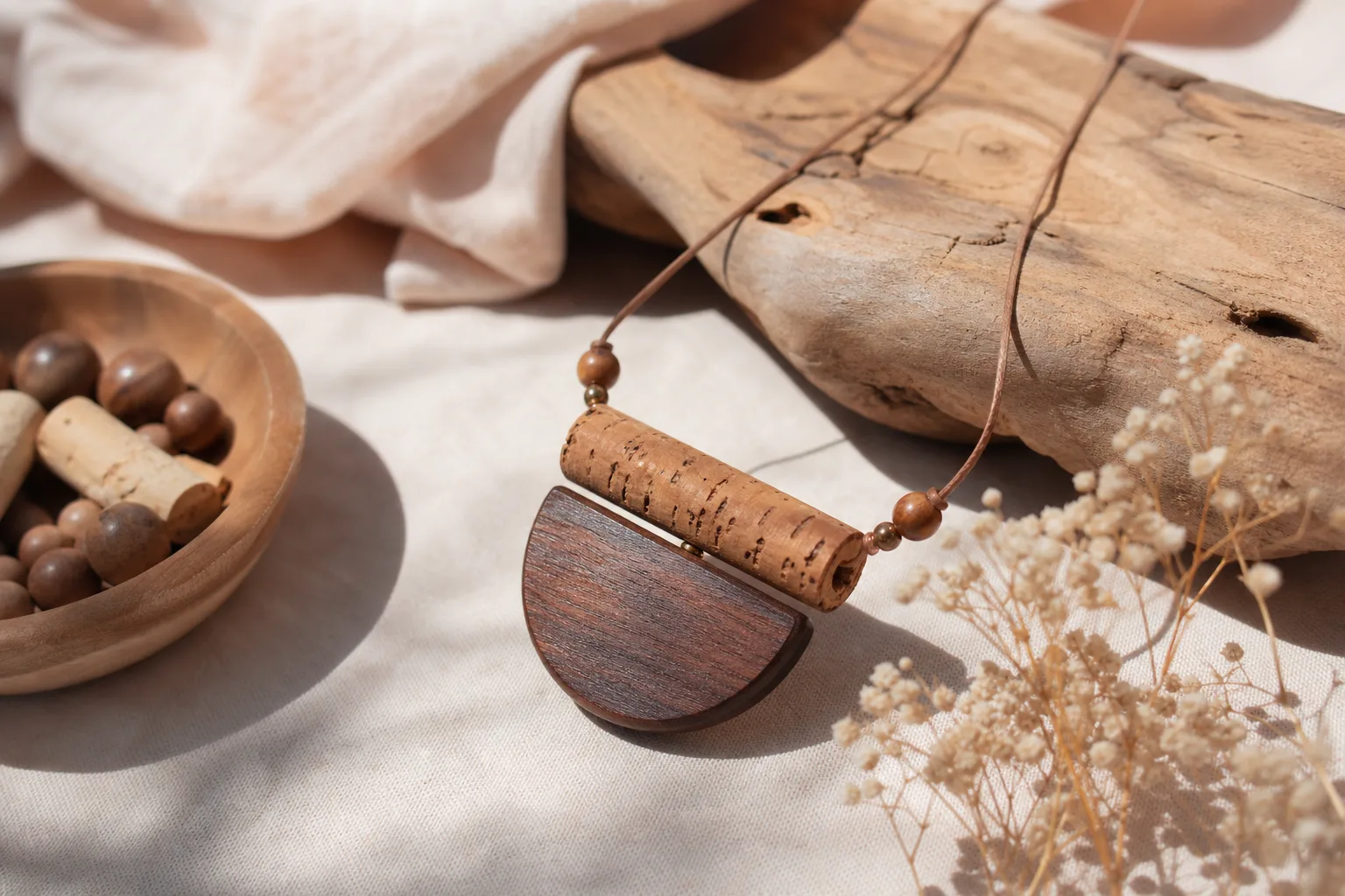Collier artisanal en perles de bois clair et pendentif en liège naturel posé sur une surface en lin.