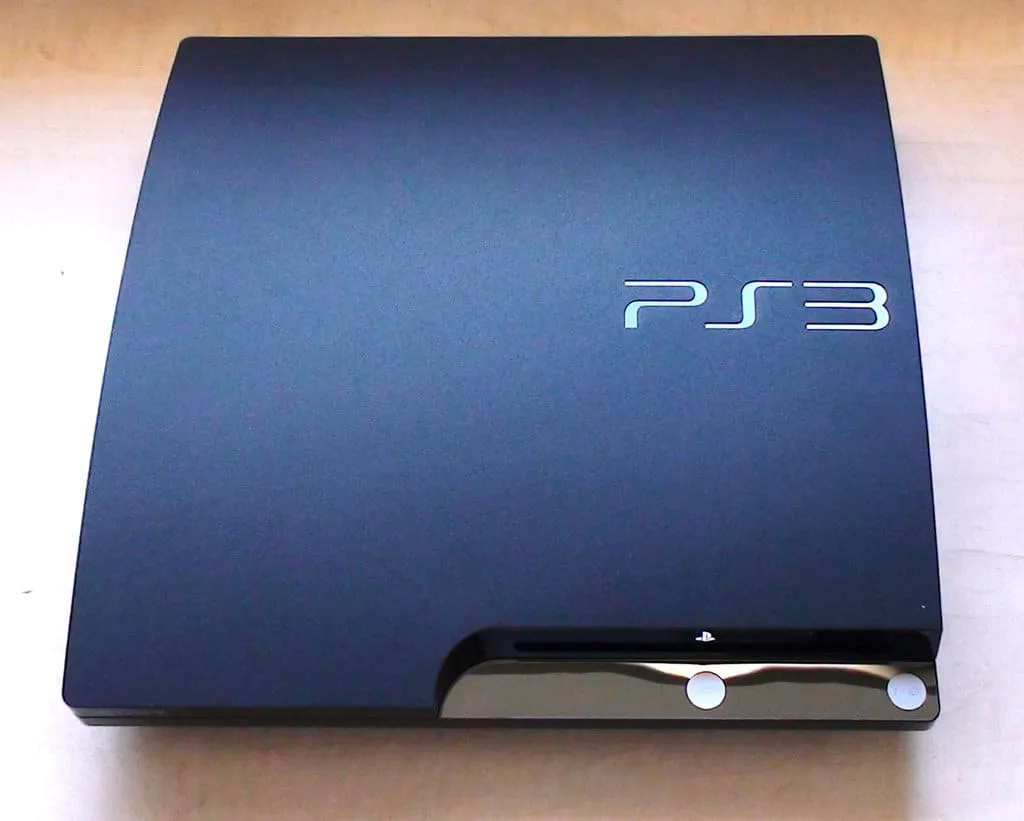 Console PlayStation 3 noire posée près de jeux vidéo, d’une manette sans fil et d’un écran de télévision.