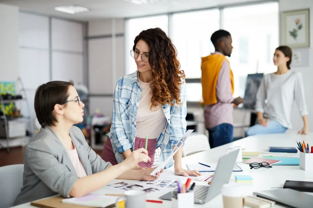 Jeune femme souriante préparant un appel téléphonique pour demander un stage, CV et carnet ouverts sur un bureau.