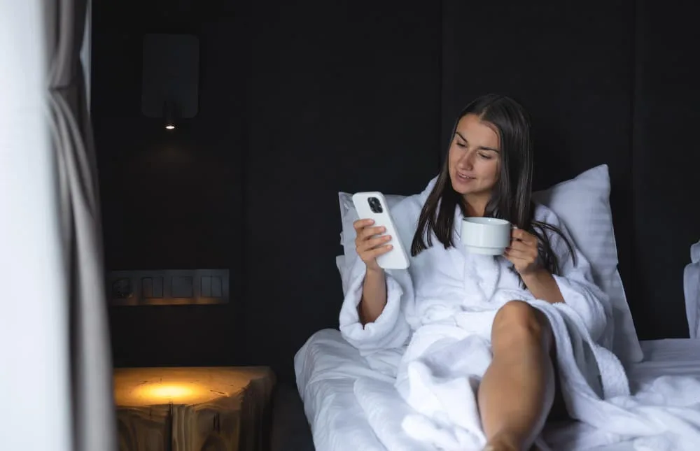 Femme assise près d’une fenêtre au lever du soleil, savourant une boisson chaude dans une ambiance matinale apaisante.
