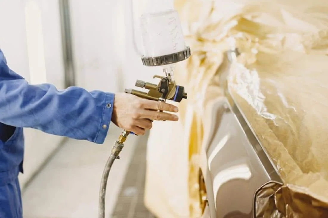 Aérosols de peinture automobile, apprêt et vernis posés près d’un pare-chocs de voiture à retoucher.
